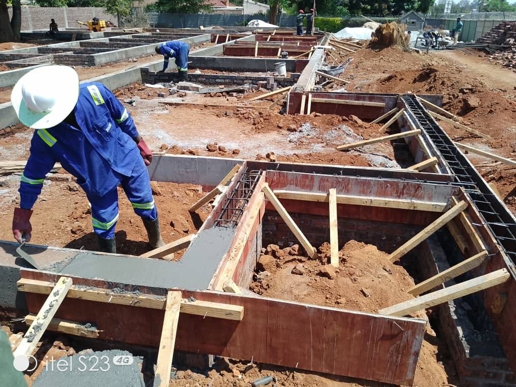 Construction workers in blue uniforms and helmets working on building foundations, using concrete and wooden frames.