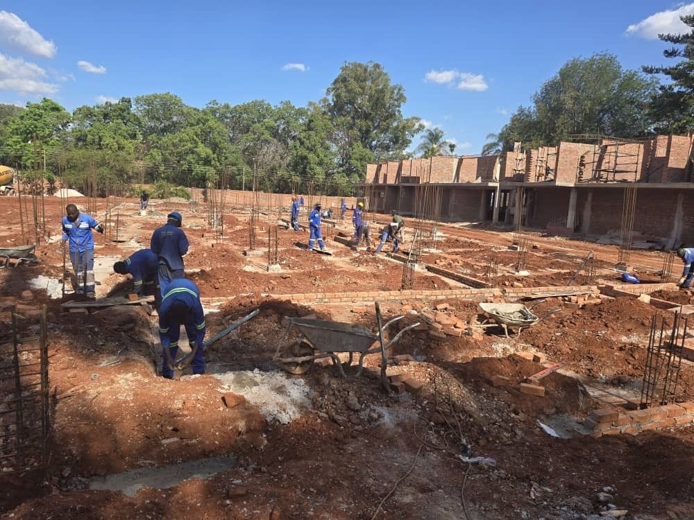 Construction site with workers laying foundations for a building, surrounded by greenery and brick structures.